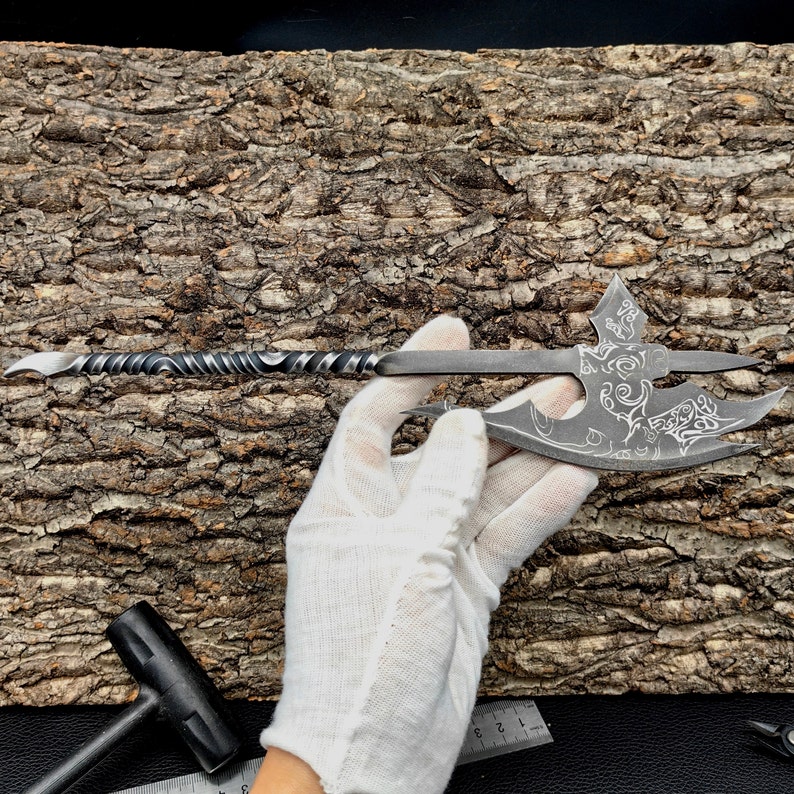 May include: A small, hand-forged axe with a twisted handle and intricate etched designs. The axe is held in a white glove against a wooden background.