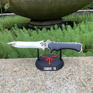 Puede incluir: Un cuchillo plateado y negro con un mango texturizado, exhibido en un soporte negro. El soporte presenta un emblema rojo y el texto "RESIDENT EVIL". El cuchillo tiene un dise&ntilde;o decorativo en la hoja.