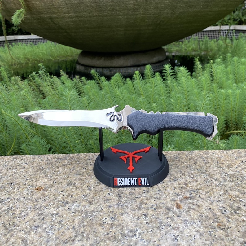 Puede incluir: Un cuchillo plateado y negro con un mango texturizado, exhibido en un soporte negro. El soporte presenta un emblema rojo y el texto "RESIDENT EVIL". El cuchillo tiene un dise&ntilde;o decorativo en la hoja.