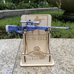 May include: A detailed toy sniper rifle in blue and white, presented on a wooden stand. The stand is engraved with "CS:GO" and a soldier silhouette. The rifle has a scope and is displayed against a natural backdrop.