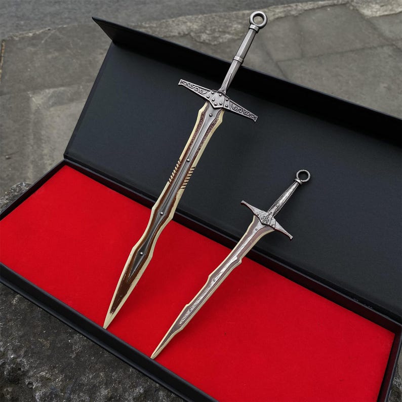 May include: Two decorative metal swords with intricate designs, one large and one small, displayed in a black box with a red interior. The swords have silver blades and handles, with detailed crossguards and pommels.