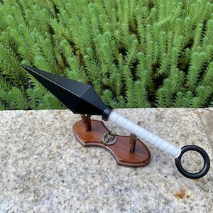 May include: A black kunai knife with a white wrapped handle and a black ring at the end. The kunai is displayed on a wooden stand with a decorative emblem. The background features green foliage.