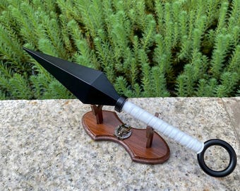 May include: A black kunai knife with a white wrapped handle and a black ring at the end. The kunai is displayed on a wooden stand with a decorative emblem. The background features green foliage.