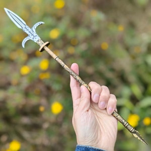 May include: A decorative spear with a silver blade and ornate gold detailing. The spearhead has a double-edged blade and curved side projections. The shaft is gold with intricate dragon and floral patterns. The spear is held against a blurred background of green and yellow.