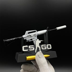 Pu&ograve; includere: Un modello bianco e nero di un fucile con silenziatore, esposto su un supporto nero con la scritta "CS GO" e la sagoma di una persona che tiene un'arma. Sul supporto c'&egrave; anche un proiettile giallo.