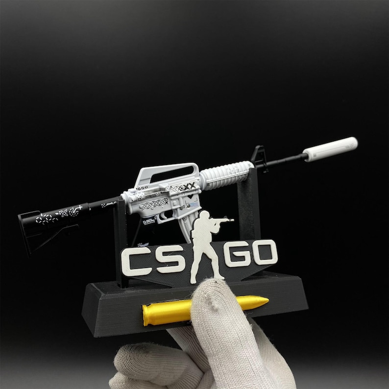 Pu&ograve; includere: Un modello bianco e nero di un fucile con silenziatore, esposto su un supporto nero con la scritta "CS GO" e la sagoma di una persona che tiene un'arma. Sul supporto c'&egrave; anche un proiettile giallo.