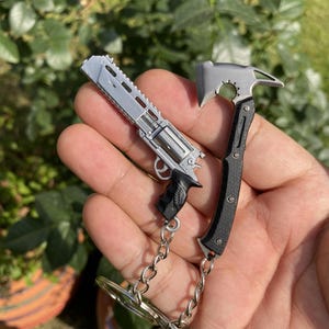 Pode incluir: Um revólver em miniatura de cor prateada com punho preto e um machado em miniatura de cor preta e prateada presos a um porta-chaves de metal. Ambos os itens apresentam designs mecânicos detalhados e estão ligados por correntes de metal.