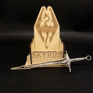 Puede incluir: Un expositor de madera con la palabra "SKYRIM" y un emblema de drag&oacute;n, sosteniendo una espada plateada. El expositor es de madera clara y la espada tiene detalles intrincados.