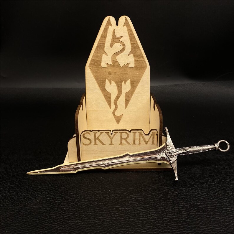 Puede incluir: Un expositor de madera con la palabra "SKYRIM" y un emblema de drag&oacute;n, sosteniendo una espada plateada. El expositor es de madera clara y la espada tiene detalles intrincados.