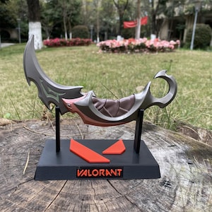 Op de afbeelding: Een grijze en bruine karambit mes replica met een gebogen lemmet, tentoongesteld op een zwarte standaard. De standaard heeft het woord "VALORANT" in witte letters en twee oranje geometrische vormen. Het mes heeft een bruin handvat en roze accenten.
