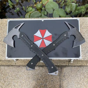May include: Two tactical tomahawks with matte grey heads and black textured handles are crossed over a black rectangular carrying case. The case features a central red and white umbrella-shaped logo with eight segments and a silver metal frame.