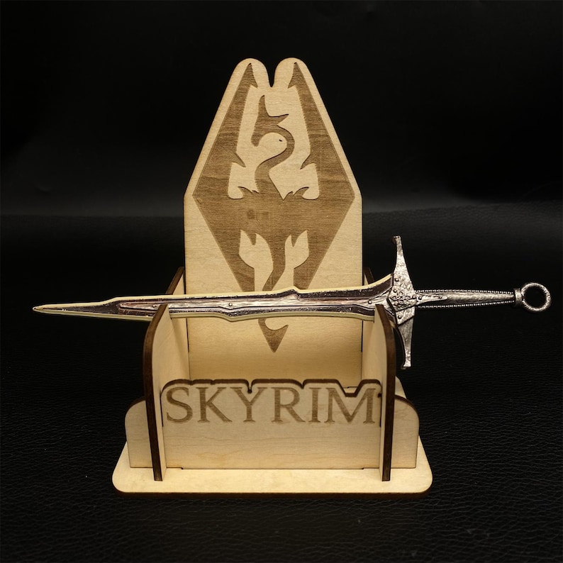 Puede incluir: Una espada plateada descansa en un soporte de madera. El soporte presenta la palabra "SKYRIM" y un emblema de drag&oacute;n. La espada tiene una empu&ntilde;adura detallada y una hoja afilada, mostrando un tema de fantas&iacute;a.