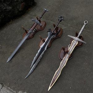 May include: Three decorative swords with ornate designs are displayed on wooden plaques. The swords feature silver blades and detailed handles. One sword has a brown handle, another has a black handle, and the third has a silver handle with a circular pommel.