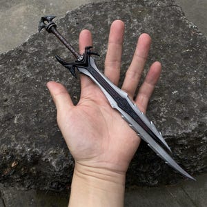 May include: A detailed, miniature sword with a black and silver blade and a brown handle. The sword features intricate designs and is held in a hand, showcasing its size and craftsmanship. The overall aesthetic suggests a fantasy or collectible item.