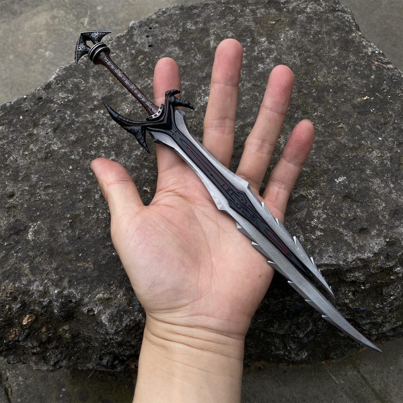 May include: A detailed, miniature sword with a black and silver blade and a brown handle. The sword features intricate designs and is held in a hand, showcasing its size and craftsmanship. The overall aesthetic suggests a fantasy or collectible item.