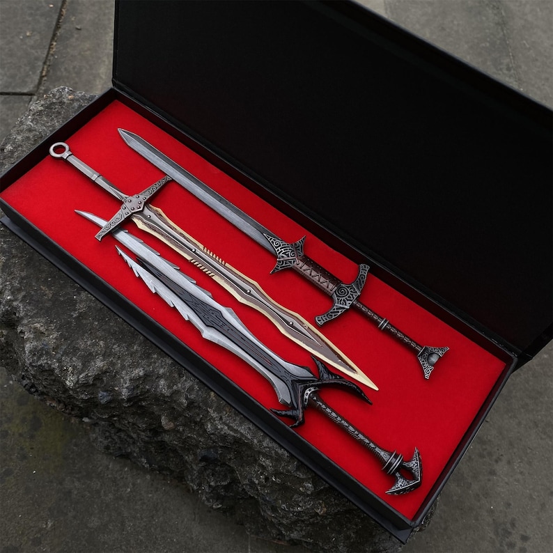 May include: A set of four decorative swords displayed in a black box with a red velvet interior. The swords have intricate designs and varying blade shapes, with silver, gold, and black finishes. The box is open, revealing the swords.