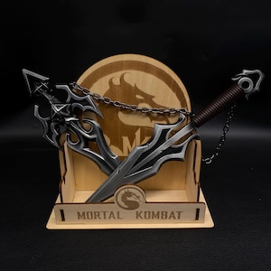 May include: A wooden display featuring two metal weapons with intricate designs, connected by a chain. The display includes the text "MORTAL KOMBAT" and a logo. The weapons have silver blades and dark accents.