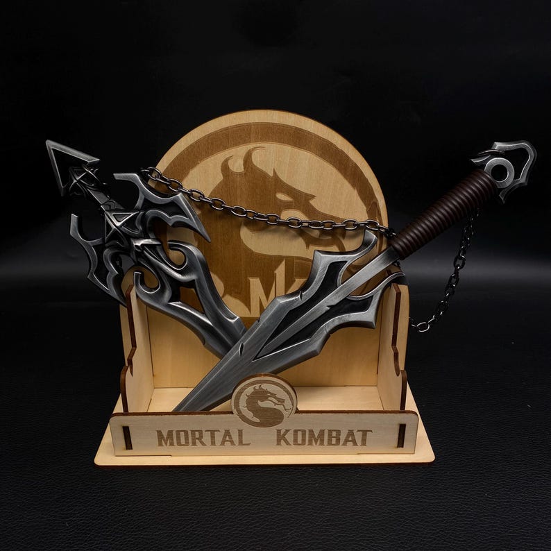 May include: A wooden display featuring two metal weapons with intricate designs, connected by a chain. The display includes the text "MORTAL KOMBAT" and a logo. The weapons have silver blades and dark accents.