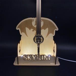May include: A wooden Skyrim-themed lamp featuring a detailed sword design. The lamp has a light-up base with the word "SKYRIM" and a silhouette of a dragon. The sword is intricately designed with a detailed hilt.