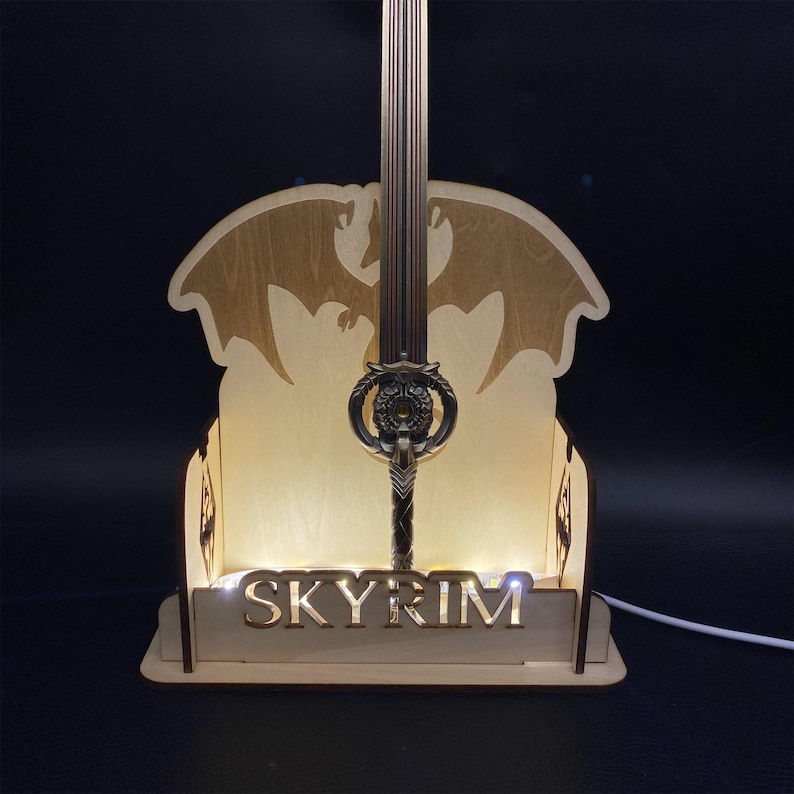 May include: A wooden Skyrim-themed lamp featuring a detailed sword design. The lamp has a light-up base with the word "SKYRIM" and a silhouette of a dragon. The sword is intricately designed with a detailed hilt.