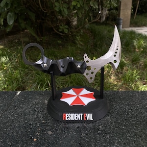 May include: A black and silver karambit knife with a curved blade and a ring handle, displayed on a black base with the Resident Evil logo. The base features a red and white umbrella design.