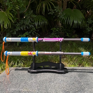 May include: Two toy weapons with colorful designs are displayed on a black stand. One has a blue and white handle, the other has a pink handle. The handles have cartoon graphics. The weapons have a silver shaft and blue and white striped ends.
