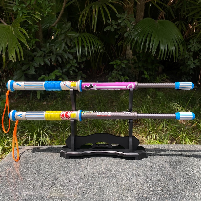 May include: Two toy weapons with colorful designs are displayed on a black stand. One has a blue and white handle, the other has a pink handle. The handles have cartoon graphics. The weapons have a silver shaft and blue and white striped ends.