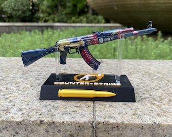 May include: A miniature replica of an AK-47 rifle featuring a multi-colored graphic pattern with blue, red, and gold accents. The model is mounted on a black display stand labeled COUNTER-STRIKE with a gold-colored bullet replica attached.
