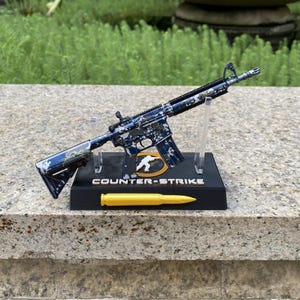 May include: A miniature rifle model featuring a blue and white camouflage pattern, mounted on a black display stand. The base includes the text COUNTER-STRIKE, a circular logo, and a yellow bullet replica positioned horizontally at the front.