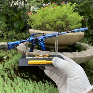 Puede incluir: Un modelo de rifle en miniatura con un patrón de llamas azules y negras, montado sobre una base negra con un detalle en forma de bala amarilla. La base incluye el texto Counter-Strike y un logotipo circular, sostenido por pilares de plástico.