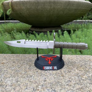 Puede incluir: Un cuchillo bayoneta plateado con borde dentado, exhibido en un soporte negro con un emblema rojo y el texto "RESIDENT EVIL". El cuchillo tiene un mango texturizado. El fondo es natural.