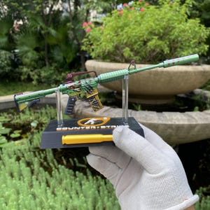 May include: A miniature replica of a rifle featuring a green and yellow camouflage pattern with a long barrel attachment. The model is mounted on a black display stand with the text COUNTER-STRIKE and a yellow bullet-shaped accent piece attached.