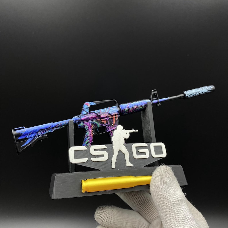 Pu&ograve; includere: Una replica in miniatura di un fucile con una finitura iridescente colorata, supportata da un supporto nero. Il supporto presenta il testo "CS GO" e una silhouette di una persona che tiene una pistola. &Egrave; presente anche un proiettile dorato.