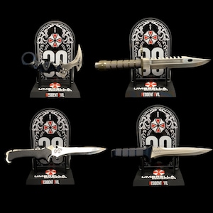 Könnte beinhalten: Vier Sammlermesser auf Präsentationsständern. Jeder Ständer zeigt das Umbrella Corporation-Logo und den Text "Resident Evil". Die Messer haben silberne Klingen und schwarze oder graue Griffe. Die Ständer sind schwarz mit weißen und roten Akzenten.