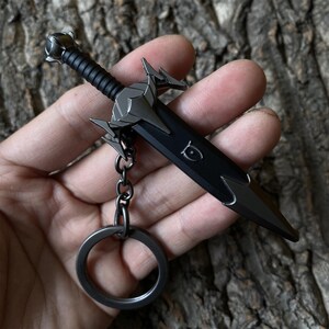 Puede incluir: Un llavero de daga negro y plateado. La espada en miniatura presenta una empu&ntilde;adura y guarda detalladas, con un mango estriado y una hoja puntiaguda. El anillo y la cadena del llavero tambi&eacute;n son plateados. La daga tiene un dise&ntilde;o decorativo en la hoja.