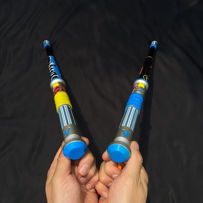 May include: Two colorful toy light sabers with blue, yellow, and silver accents. The handles are held in hands against a black background. The light sabers have a blue tip and a silver base.