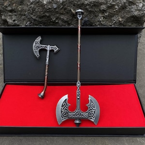 May include: Two miniature axes presented in a black box with a red interior. The axes feature silver-coloured blades with detailed patterns and brown handles. One is a small, single-bladed axe, and the other is a larger, double-bladed axe.