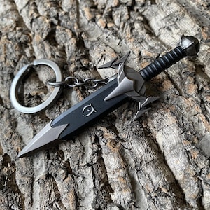 Puede incluir: Un llavero de espada en miniatura con una hoja negra y detalles plateados. El mango es negro con un agarre texturizado, y el llavero incluye un anillo y una cadena plateados. La espada descansa sobre una superficie de madera marr&oacute;n texturizada.