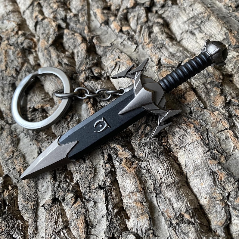 Puede incluir: Un llavero de espada en miniatura con una hoja negra y detalles plateados. El mango es negro con un agarre texturizado, y el llavero incluye un anillo y una cadena plateados. La espada descansa sobre una superficie de madera marr&oacute;n texturizada.