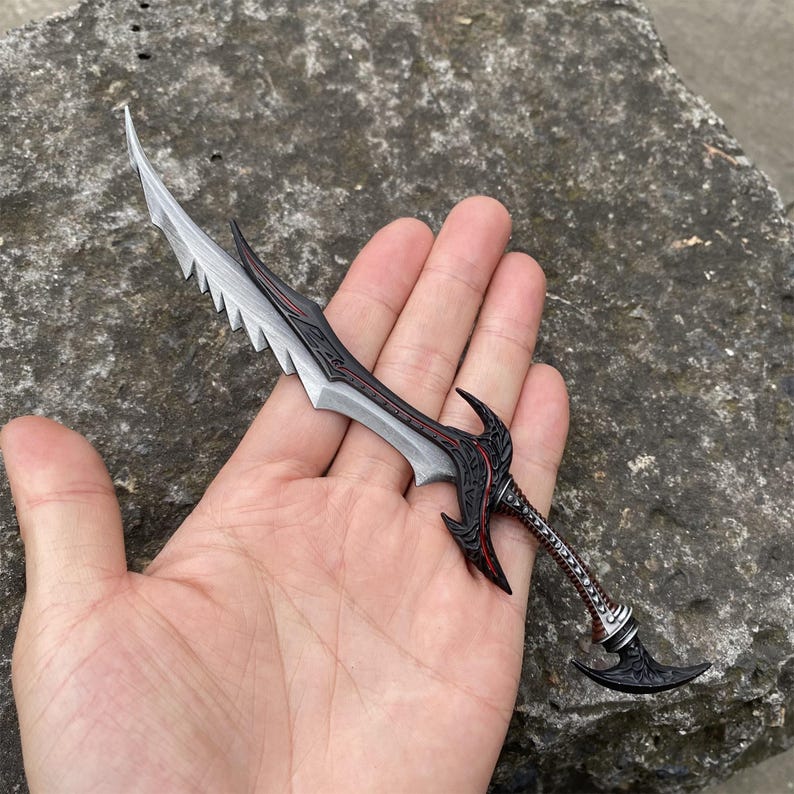 May include: A detailed miniature sword with a silver blade and black handle, accented with red detailing. The blade has a serrated edge. The handle features intricate patterns and a curved pommel. The sword is held in a hand.