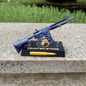 May include: A miniature rifle model featuring a blue and white marbled pattern, mounted on a black display stand with the text Counter-Strike and a circular logo. A single gold-coloured bullet replica rests horizontally on the base in front.