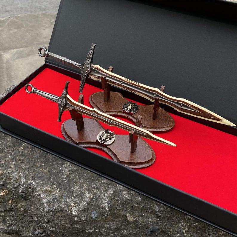 May include: Two decorative swords with ornate handles and blades, displayed in a black box with a red interior. The swords are mounted on wooden stands with decorative medallions. The blades have a gold and brown color scheme.