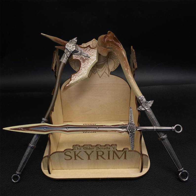 May include: A wooden display stand with the text "SKYRIM" holds a collection of fantasy weapons. The weapons include a silver and brown axe, a curved bone-like weapon, and a long sword with a silver handle and blade.