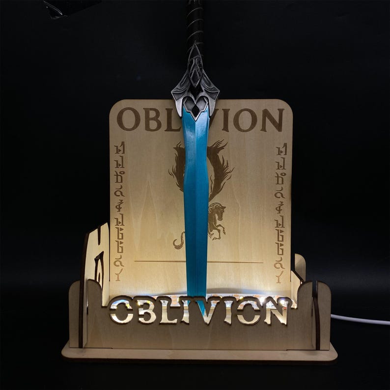 May include: A wooden display stand with the word "OBLIVION" and a blue-bladed sword. The stand has a light-colored wooden finish with the word "OBLIVION" cut out at the base. The sword has a decorative hilt.