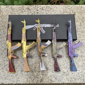 May include: Five miniature rifle models arranged in a row on a black rectangular box. The models feature distinct finishes including wood grain, gold with intricate patterns, metallic purple, and a leopard print design with a dark stock.