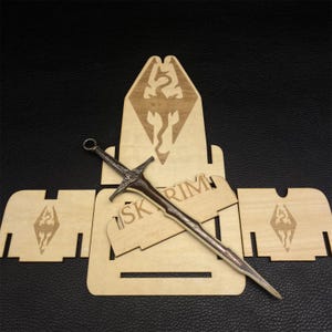 Puede incluir: Una espada de metal detallada con un mango marr&oacute;n y una hoja plateada descansa sobre un soporte de exhibici&oacute;n de madera. El soporte presenta la palabra "SKYRIM" y un emblema de drag&oacute;n. Tambi&eacute;n hay piezas de madera adicionales con el mismo emblema.