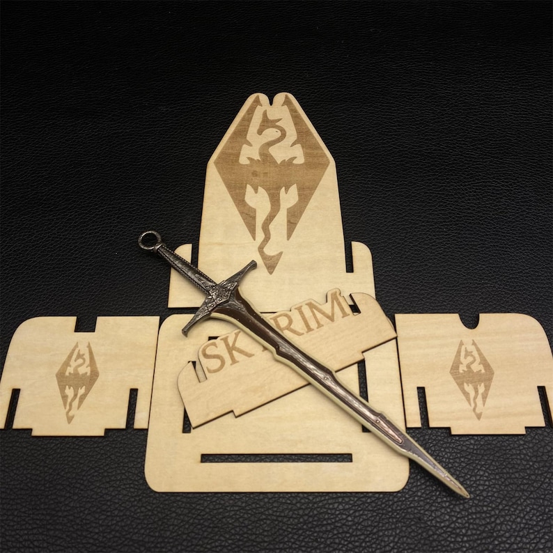 Puede incluir: Una espada de metal detallada con un mango marr&oacute;n y una hoja plateada descansa sobre un soporte de exhibici&oacute;n de madera. El soporte presenta la palabra "SKYRIM" y un emblema de drag&oacute;n. Tambi&eacute;n hay piezas de madera adicionales con el mismo emblema.