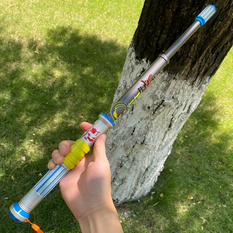 May include: A multi-colored toy sword with a silver shaft, blue and yellow accents, and a red emblem. The toy has a blue tip and an orange tassel. The sword is held in a hand against a background of green grass and a tree trunk.