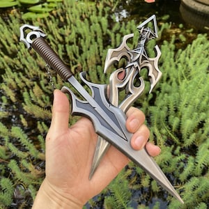 May include: Two decorative daggers with ornate, silver-colored blades and handles. One dagger has a brown, wrapped handle and a chain. The other dagger has a decorative hilt with a triangular design. The daggers are held against a backdrop of green foliage.