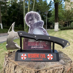 May include: A silver and black axe with a textured handle, displayed on a black stand. The stand features the text "RESIDENT EVIL" and a red and white umbrella logo. A cardboard cutout of a character is in the background.